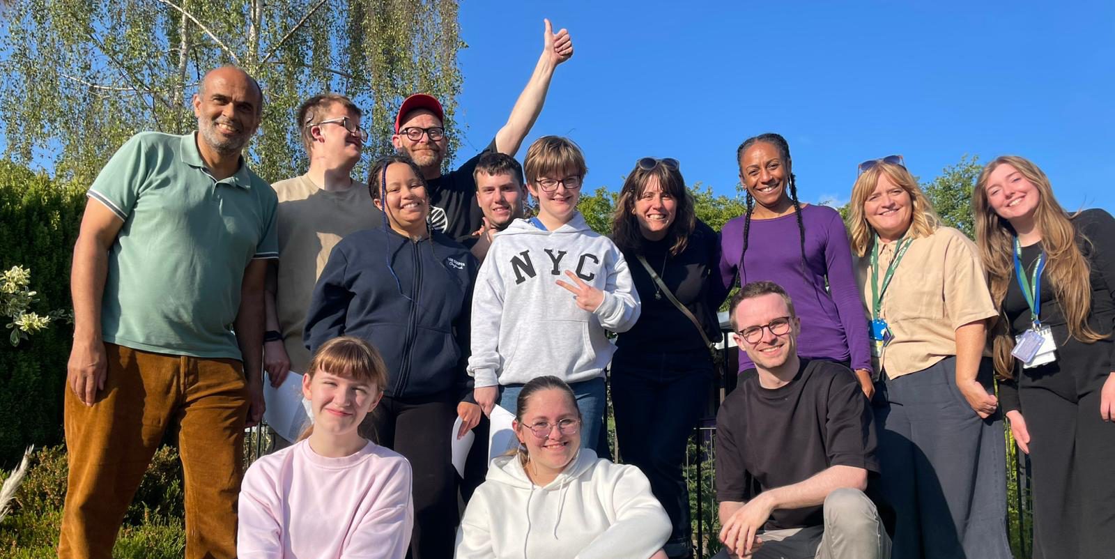 A group of 13 individuals smiling into the camera. A clear blue sky and trees in the background